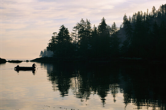 Silhouetted boater at twilight near tree-lined shoreline; Ramsay Island, Queen Charlotte Islands, British Columbia, Canada