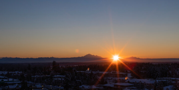 Sunburst over mountains and cityscape at sunset; Surrey, British Columbia, Canada