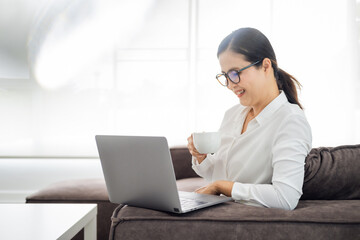 Enjoying time at home. Beautiful smiling woman working on laptop and drinking coffee while sitting in a big comfortable chair at home