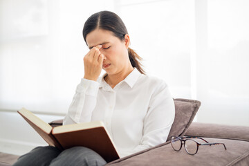 Lady suffering eyestrain reading book sitting on a couch in living room at home