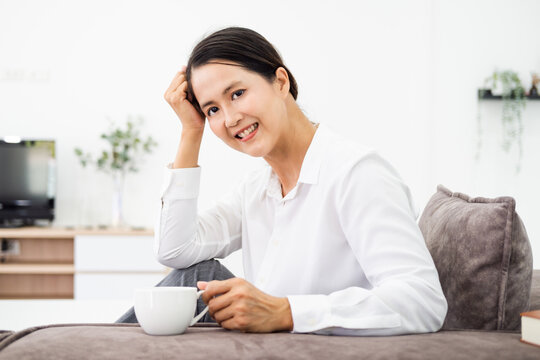 Happy beautiful relaxed mature  adult asian woman drinking coffee relaxing on sofa at home. Smiling stylish middle aged 40s lady enjoying resting sitting on couch in modern living room.