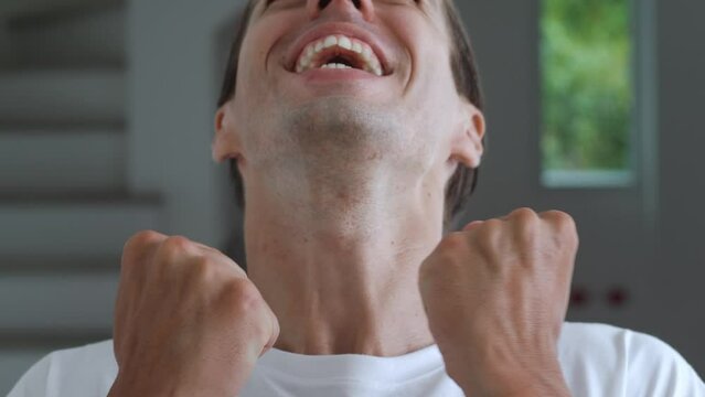 Close up portrait of happy young man 30s wears white t-shirt look camera celebrate win scream rejoices doing winner hands gesture isolated standing at home. Male winner clenching fists say yes.