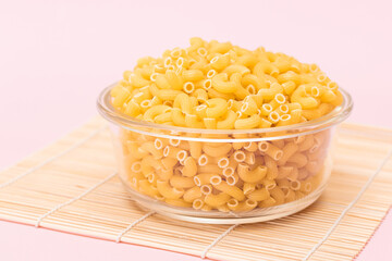 Uncooked Chifferi Rigati Pasta in Glass Jar on Bamboo Mat on Pink Background. Fat and Unhealthy Food. Classic Dry Macaroni. Italian Culture and Cuisine. Raw Pasta