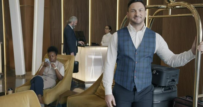 Cheerful bellboy wearing uniform is standing in hotel lobby holding luggage cart and smiling looking at camera. Occupation and people concept.