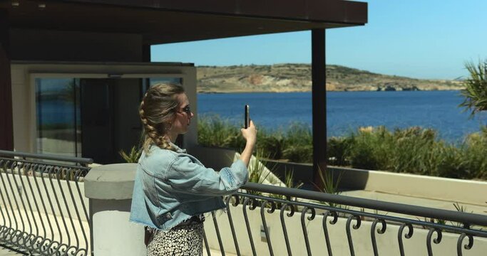 Young stylish woman at seafront shoots video of sea area landscape on smartphone. Middle age woman in denim jacket and dress creates story or reel for social media on sunny day.