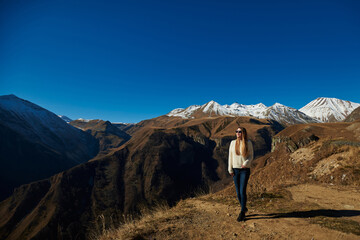 a beautiful young woman in sunglasses with long hair in a light sweater and jeans against the backdrop of sharp snowy peaks of high mountains. vacation in the mountains. trip to the mountains