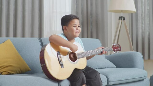 Talented Asian Kid Practicing Singing And Playing The Guitar On Sofa At Home
