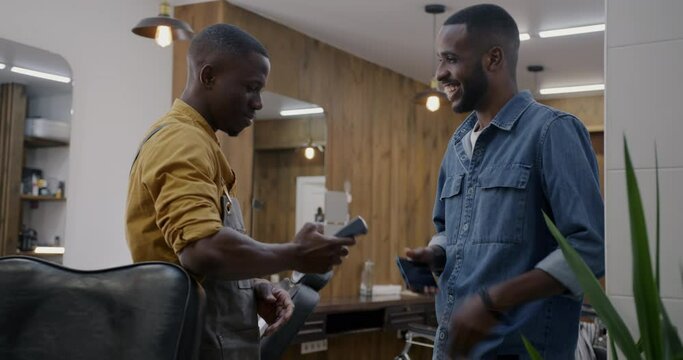 Barber accepting smartphone payment using modern terminal with wireless technology and shaking hands with client in barbershop. People and hairdressing service concept.