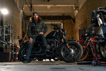 Obraz na płótnie Canvas Creative authentic motorcycle workshop garage serious redhead bearded biker mechanic sitting on motorcycle