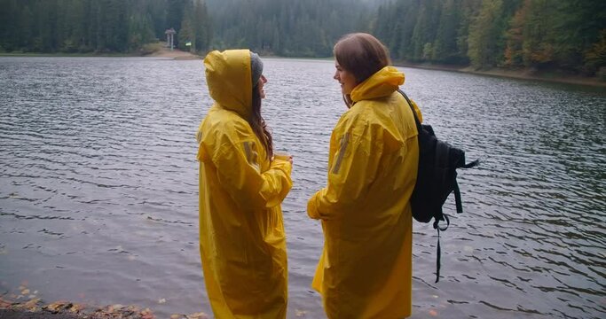 Two happy women talking standing on coast of lake Synevyr smiling. Caucasian female tourists enjoying vacations in Carpathian mountains in Ukraine. Cinema 4k ProRes HQ