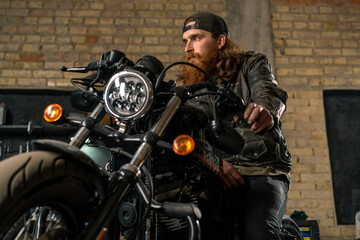 Fototapeta na wymiar Creative authentic motorcycle workshop garage portrait serious redhead bearded biker mechanic sitting on motorcycle