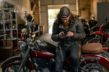 Fototapeta na wymiar Creative authentic motorcycle workshop Garage brutal serious bearded redhead biker mechanic sitting on motorcycle with phone in hands