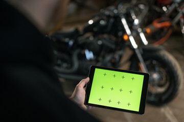 Creative authentic motorcycle workshop Biker mechanic in garage standing near motorcycle and holding green screen tablet