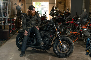 Plakat Creative authentic motorcycle workshop garage serious redhead bearded biker mechanic sitting on motorcycle