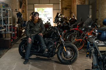 Naklejka na ściany i meble Creative authentic motorcycle workshop garage serious redhead bearded biker mechanic sitting on motorcycle