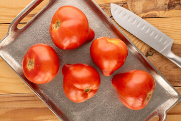 Several red tomatoes with metal tray and knife on wooden table, macro, top view.