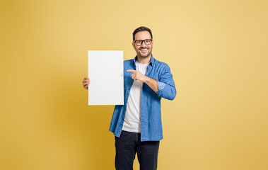Smiling businessman pointing at blank white placard for advertising against yellow background