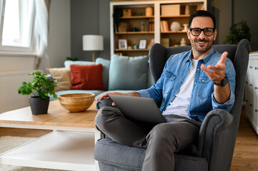 Happy businessman with laptop gesturing at camera while sitting on armchair in home office