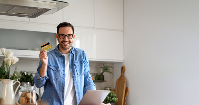 Portrait Of Cheerful Businessman Showing Credit Card And Using Laptop While Standing In Home Office