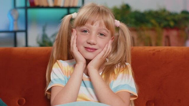 Close-up portrait of happy smiling Caucasian preteen school girl 7 years old. Young blonde lovely child kid looking at camera at home play room apartment sitting on orange couch. Ponytails hairstyle