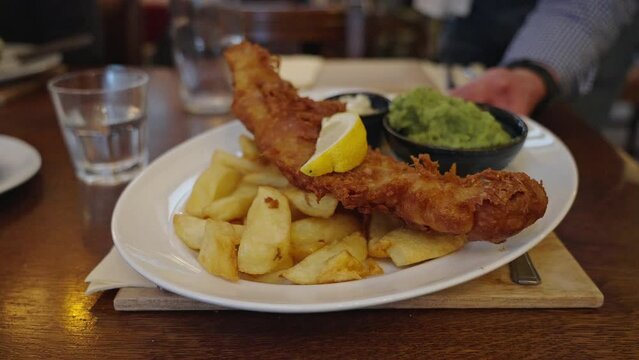 A server places a plate of traditional fish and chips with mushy peas on a table in a restaurant in Conwy, Wales, UK