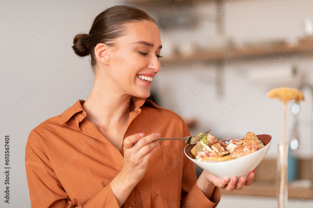 Wall mural satisfied happy young european woman enjoying tasty caesar salad, sitting in kitchen interior at hom