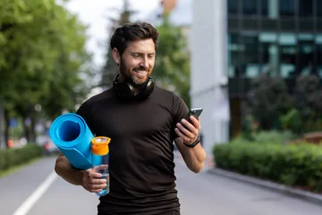 Gordijnen Portrait of mature successful adult athlete, bearded man going to workout, coach holding fitness mat and phone, browsing online app for joint physical training © Tetiana