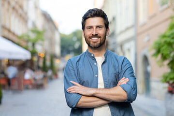 Portrait of a mature successful man in the city, the guy is smiling and looking at the camera with his arms crossed, walking in the evening city on a trip