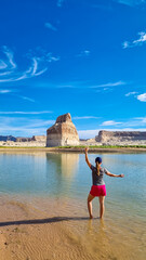 Fototapeta na wymiar Rear view of woman walking at lakeshore looking at solitary rock formation Lone Rock in Wahweap Bay at Lake Powell, Glen Canyon Recreation Area, Page, Utah, USA. Lone rock campground in sunny summer