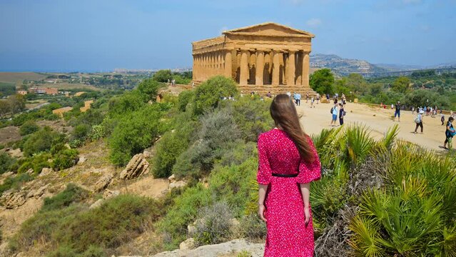 A beautiful woman in a red dress visits the Temple of Concordia in Agrigento. Ancient, preserved Greek temple on a hill, with Doric architecture and historical constructions in Sicily, Italy