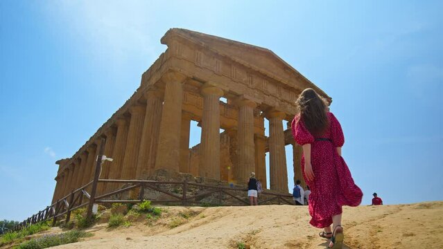 A beautiful woman in a red dress visits the Temple of Concordia in Agrigento. Ancient, preserved Greek temple on a hill, with Doric architecture and historical constructions in Sicily, Italy.
