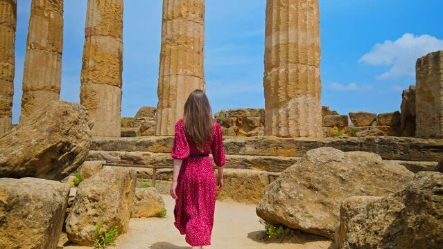 A female tourist visits the antique Temple of Heracles in Sicily, Italy. A young woman in a beautiful long dress enjoys the view of the greek majestic temple with massive columns in Agrigento.