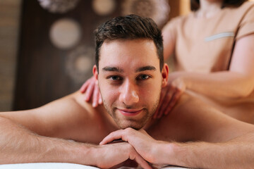 Fototapeta na wymiar Portrait of happy young man relaxing with oil massage on back at spa resort looking at camera. Closeup face of handsome male receiving back massage during body treatment. Concept of healthy lifestyle.