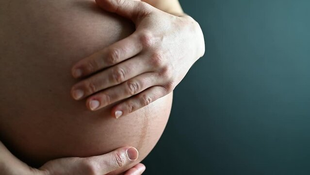 Closeup of naked round belly caressed by hands of pregnant mother in underwear. Last month of pregnancy - week 36. Side angle view. Turqouise background.