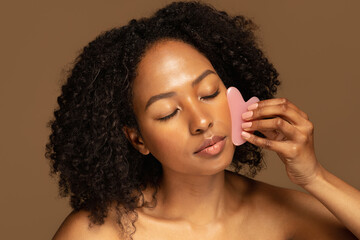 Joyful pretty african woman using gua sha stone, massaging face