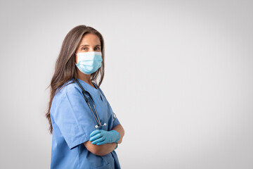 Professional female doctor in protective face mask, posing with folded arms looking at camera over...