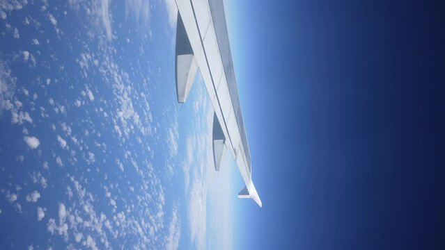 Vertical view through window on wing of plane flying above white bright clouds on background of blue sky on sunny day, slow motion. Slowly moving fluffy clouds from airplane window during flight.
