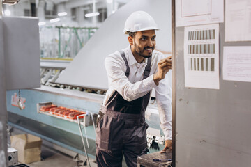 Operator of machine. Industrial worker indoors in factory. Young technician in white hard hat.