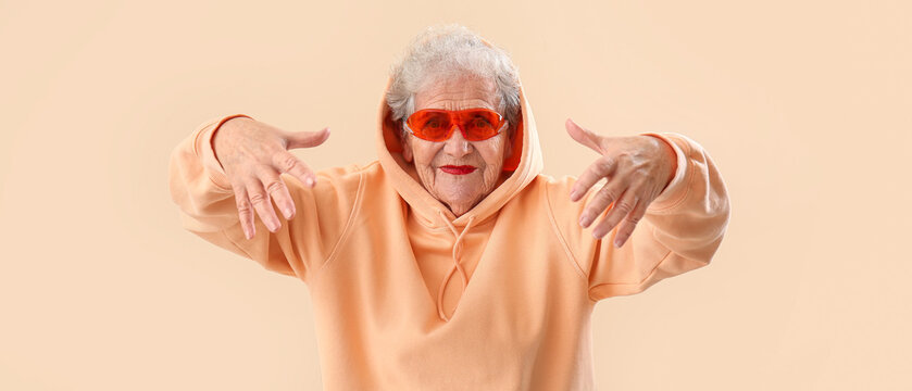 Stylish senior woman in sunglasses and hoodie on beige background
