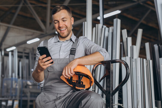 Portrait of a happy worker in a orange helmet and overalls holding a hydraulic truck and talking on the phone against a background of a factory and aluminum frames. - Powered by Adobe
