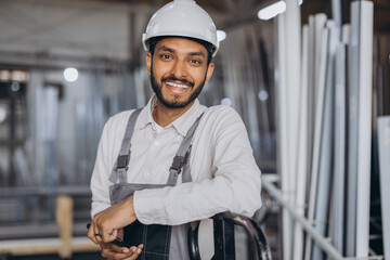 Portrait of a happy Hindu worker in a white hard hat and overalls holding a hydraulic truck against...
