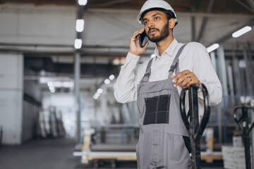 Portrait of a happy Hindu worker in a white helmet and overalls holding a hydraulic truck and...