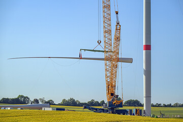 Wind turbine construction site, crane is lifting a blade to install it onto the tower, heavy industry for electricity, renewable energy and power, rural landscape, blue sky, copy space, selected focus - 615596050