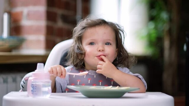 portrait caucasian baby girl about 2 years old in bib eating pasta from plate sitting high chair, self feeding for kids, baby-led weaning idea, healthy nutrition of solid food for infant