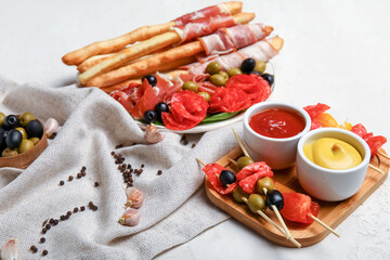 Wooden board of tasty skewers with sausages, tomatoes, olives and sauces on light background