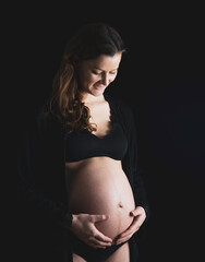 Atmospheric shot of pregnant woman in underwear gently holding her naked round belly. Last month of pregnancy - 36th week. Side angle view. Black background.