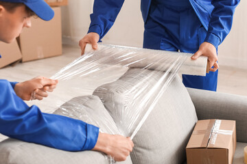 Male movers wrapping sofa with stretch film in room, closeup
