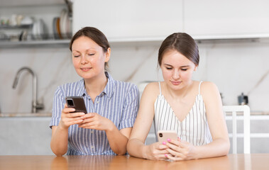 Interested middle-aged mother and teen daughter looking at their phones sitting at table in the...