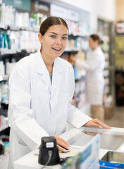 Young female pharmacist in medical uniform posing at cash register while working in pharmacy