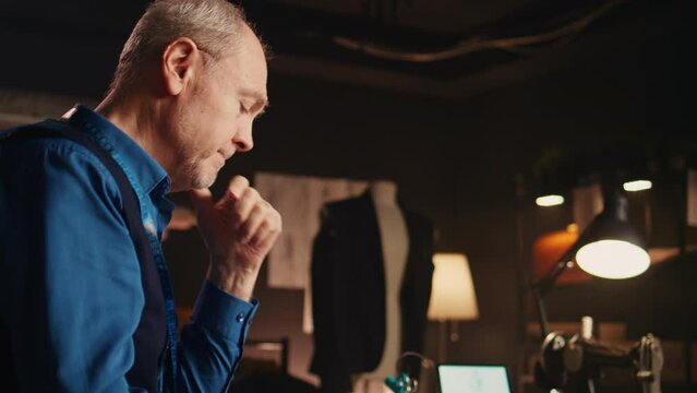 Old tailor working under pressure, feeling stressed about fashion collection ideas. Professional craftsman trying to design luxury items, frustrated man failing to find fabric. Handheld shot.